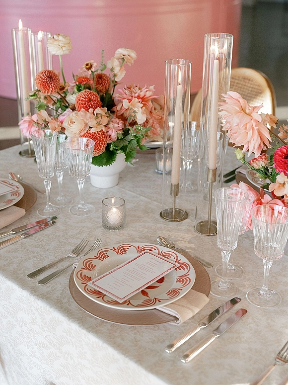Reception tablescape with wedding table centerpiece of dahlias and roses, taper candles in glass holders, patterned plates and menu card against a pink wall