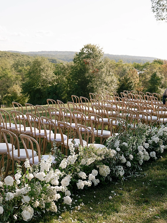 Ceremony aisle design with outdoor ceremony aisle flowers, wood chairs with white cushions, and a grounded white rose garland on a lawn