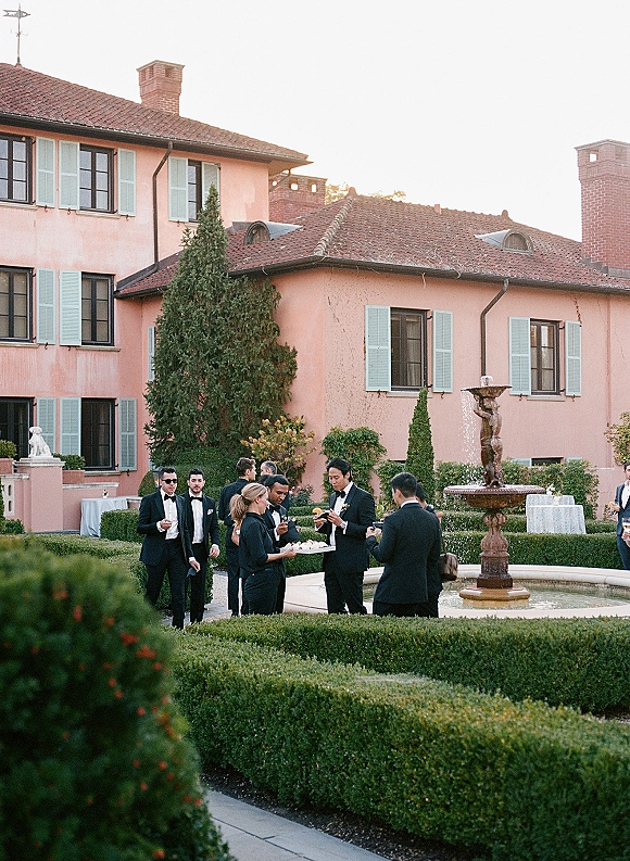 Cocktail hour wedding cocktail hour as guests in tuxedos and bow ties mingle with champagne glasses by a villa courtyard fountain