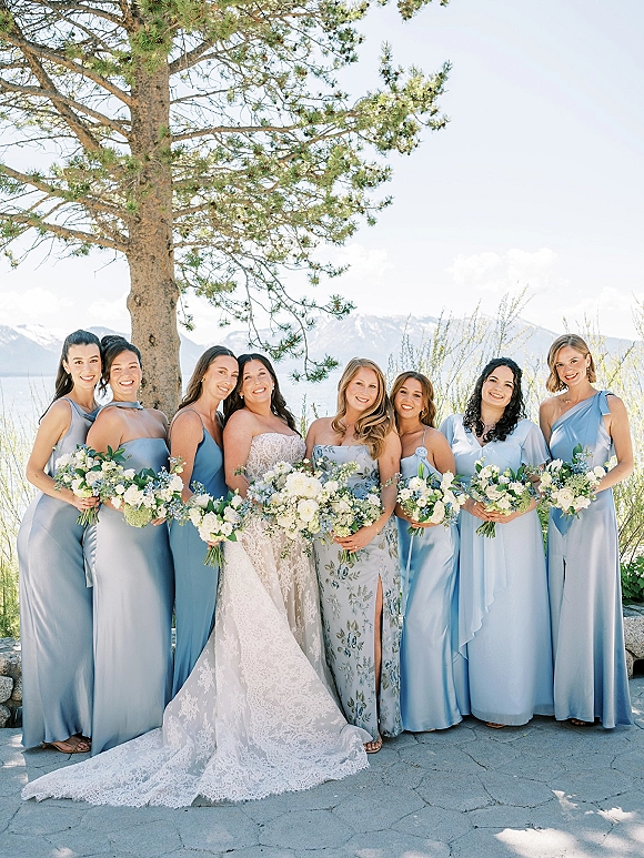 Bridesmaid group photo with bride and blue bridesmaid dresses holding white and green bouquets on a lakeside patio with mountains beyond