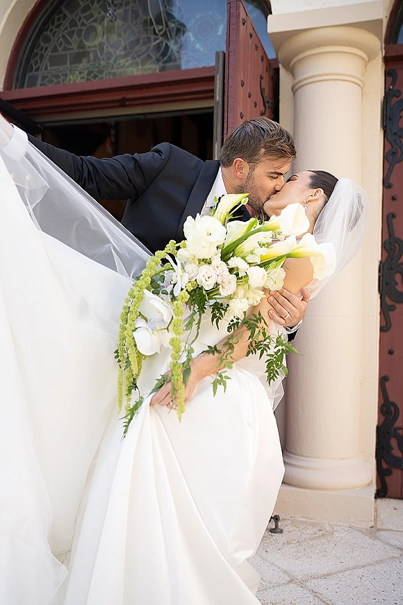 Wedding kiss portrait of groom dipping the bride as her veil sweeps and she holds a calla lily bouquet at an arched church doorway