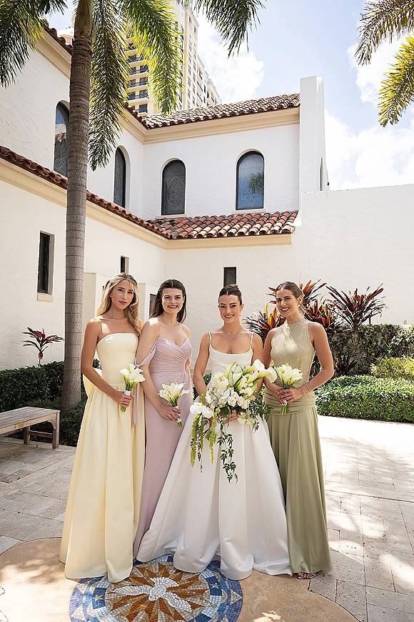 Bridesmaid group photo with bride and bridesmaids holding calla lily bouquets in a sunny courtyard by a white stucco building