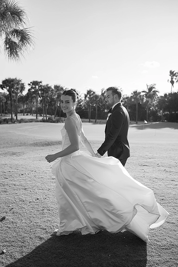 Couple portrait of bride and groom holding hands, walking on a golf course lawn with palm trees, her dress train flowing behind