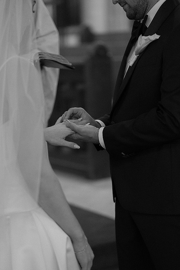 Wedding ring exchange as the groom puts a band on the bride’s hand, her veil visible, with officiant and blurred guests in a church interior