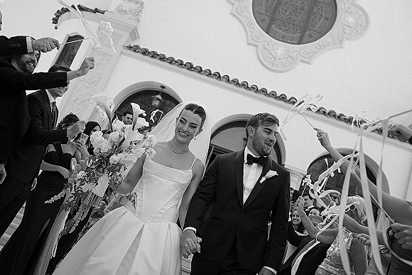 Wedding recessional as bride and groom exit a church doorway holding hands, she carries a bouquet while guests wave ribbon wands on the steps