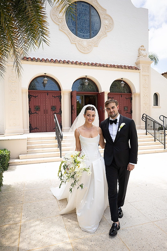 Couple portrait of bride in veil holding a calla lily bouquet beside groom in tuxedo on church steps with red doors and arches