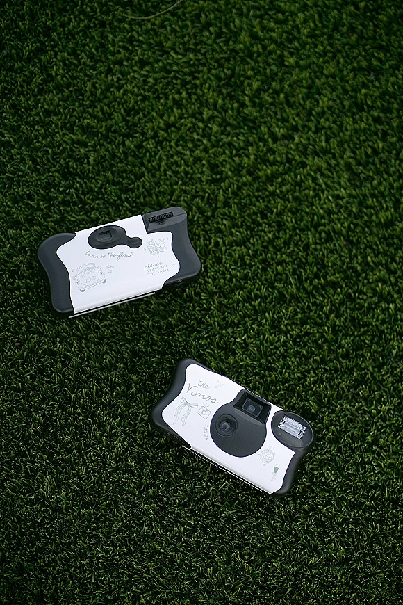 Disposable wedding cameras with custom labels and table camera instructions card arranged on a green grass lawn for guest photos