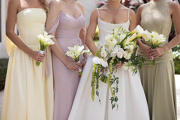Bridesmaid dresses in pastel bridesmaid dresses hues as attendants hold calla lily bouquets in sunlight beside a garden wall outdoors