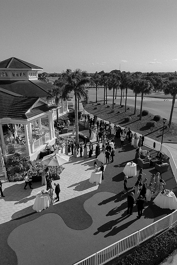 Wedding cocktail hour on a clubhouse patio with high-top tables, white linens, umbrellas, lounge seating, drinks, and palm trees by the golf course