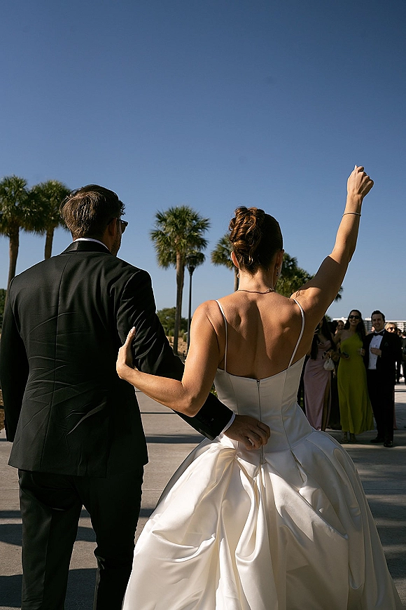 Wedding recessional as bride and groom walk away, bride raising arm in strappy back gown, guests lining palm-tree walkway under blue sky