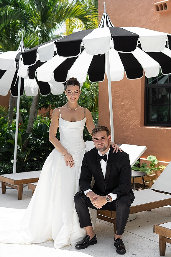 Couple portrait of bride in a white satin wedding dress beside groom in tuxedo under a striped patio umbrella with palm trees behind
