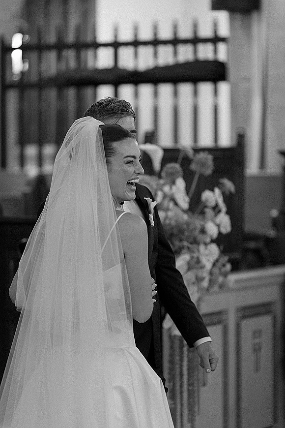 Wedding couple portrait in a black and white wedding portrait as bride laughs beside groom in tuxedo, walking church aisle past wooden pews