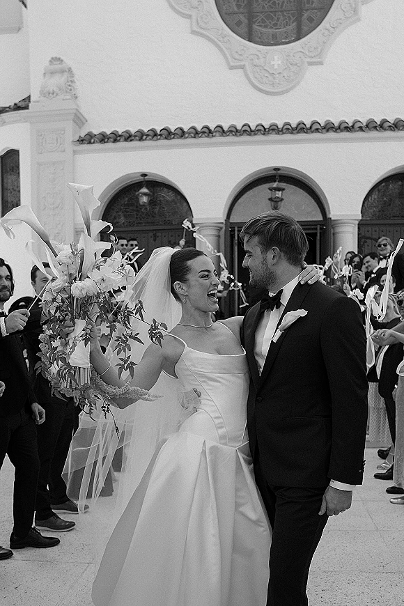 Wedding recessional as bride and groom exit the church courtyard, bride holding a calla lily bouquet while guests wave ribbon wands