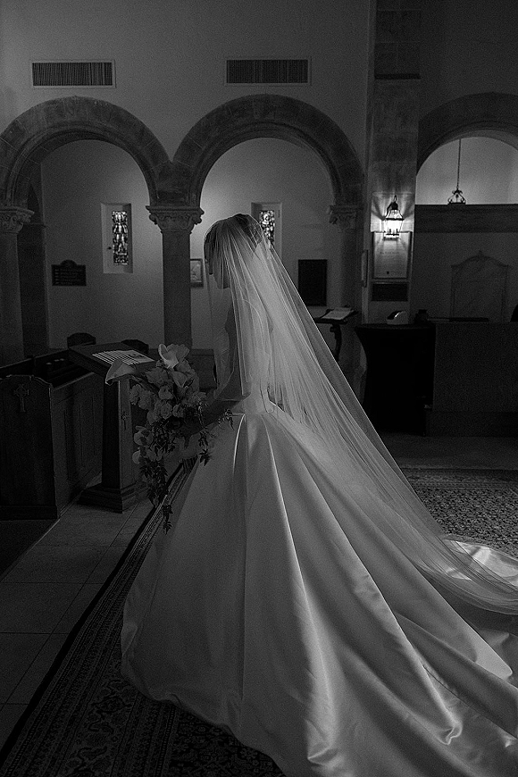 Bridal portrait of a bride from behind with a long veil and bouquet, walking down a church aisle framed by arches and stained glass windows