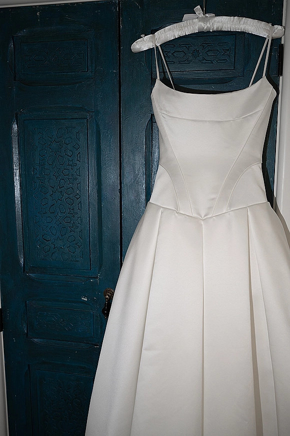 Wedding dress in simple satin wedding dress style hanging on a padded hanger with a ribbon bow against a carved wooden door