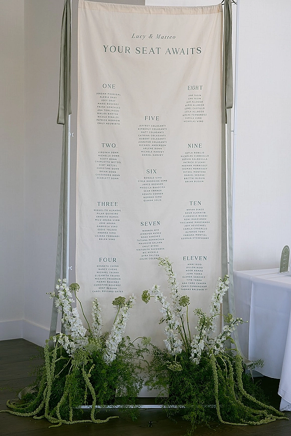 Wedding seating chart on a hanging frame with fabric seating chart banner, sage green draping, and white flowers against a white wall
