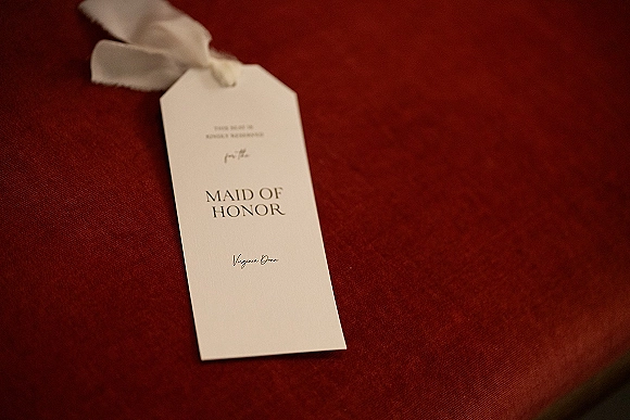 Maid of honor card with printed typography and ivory ribbon bow on a red fabric surface, styled as a wedding party proposal tag
