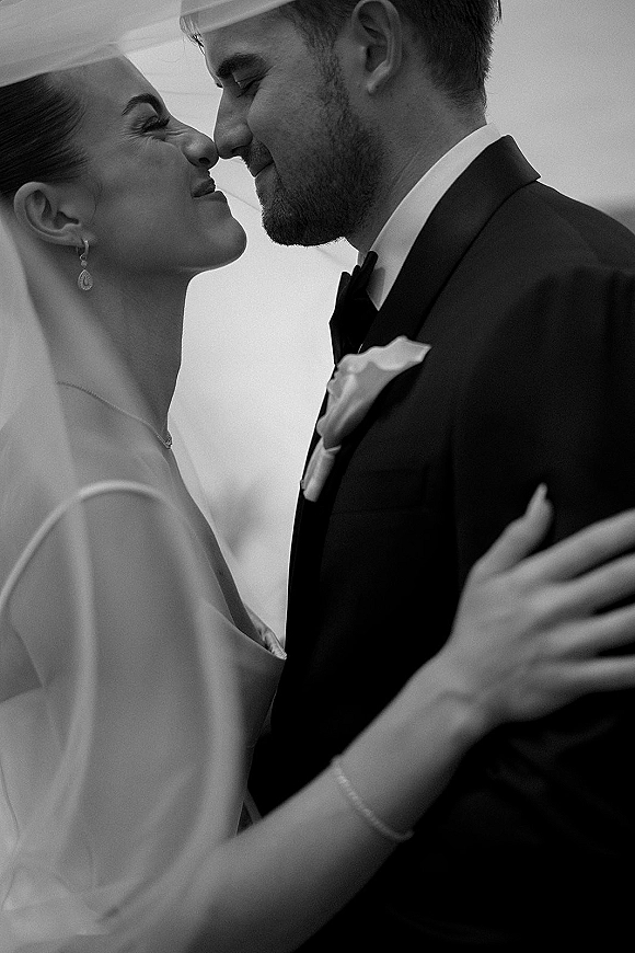 Wedding kiss portrait close up wedding kiss as bride and groom embrace, her veil draped over shoulders and his tuxedo boutonniere against a soft indoor backdrop