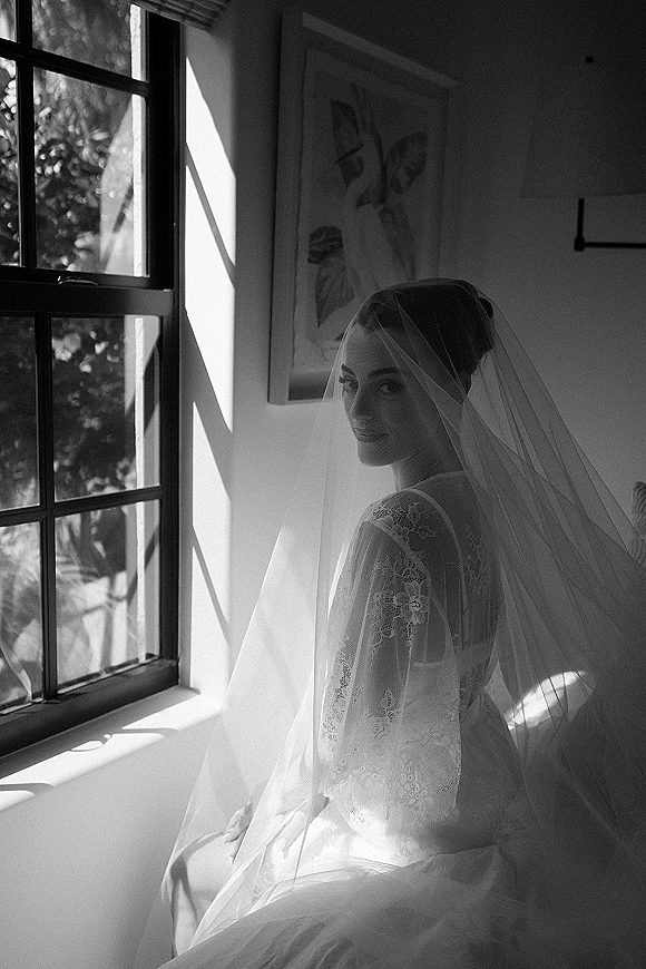 Bridal portrait in black and white of a bride by window, sheer veil over her face, lace long-sleeve dress, soft sunlight shadows