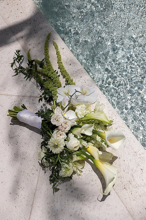 Bridal bouquet of white calla lilies with orchids and roses, wrapped in white ribbon, resting poolside on a sunlit stone deck