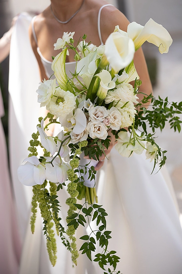 Bridal bouquet of white calla lilies, white roses, and orchids with cascading greenery held by a bride in a spaghetti-strap gown outdoors