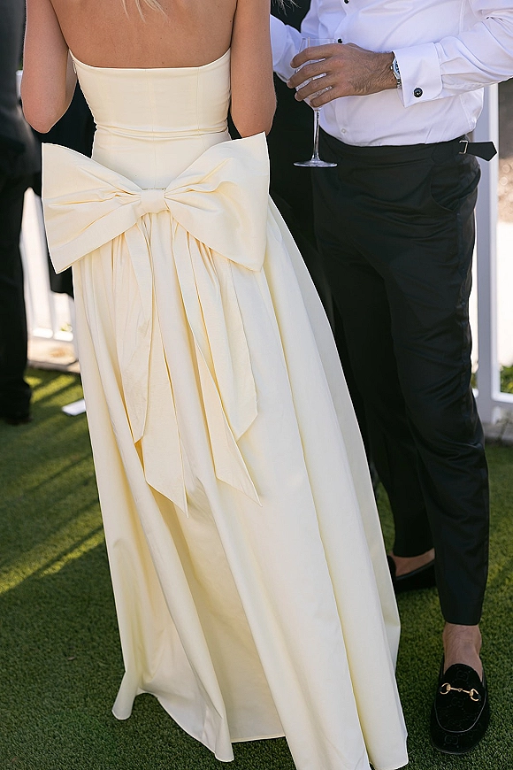 Wedding guest outfit featuring a strapless midi dress with oversized bow back and a man in black trousers, holding wine on a grass lawn