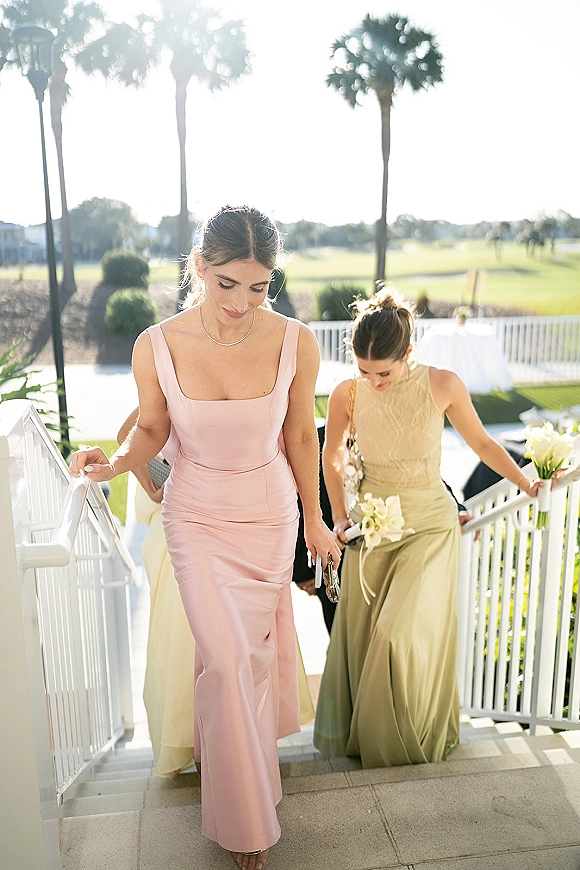 Bridesmaid dresses in blush pink and sage green as bridesmaids walk up an outdoor staircase, holding white calla lily bouquets