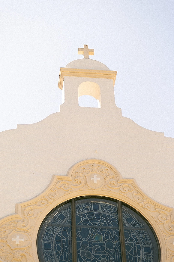 Church exterior with a white church facade and bell tower, featuring a cross and stained glass window under bright sunlight and blue sky
