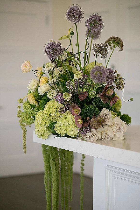 Wedding floral arrangement with tall allium, hydrangea, white roses, and hanging amaranthus on a white countertop indoors
