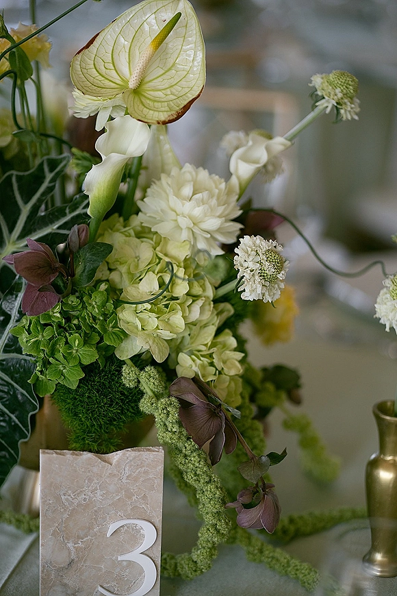 Wedding centerpiece with anthurium and calla lilies, hydrangea and greenery, plus table number card in stone holder on blurred reception table setting