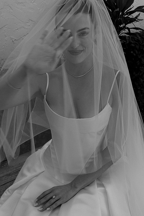 Bridal portrait of a bride under veil in a satin spaghetti-strap dress, seated by a stucco wall with greenery, showing jewelry and ring