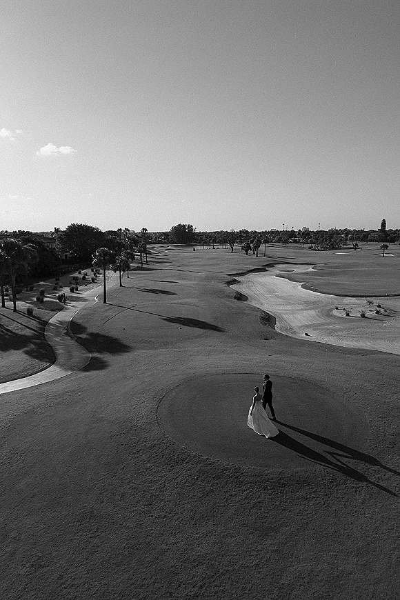 Couple portrait of bride and groom walking hand in hand, bride in wedding dress and groom in suit, across a golf course with palm trees and sky