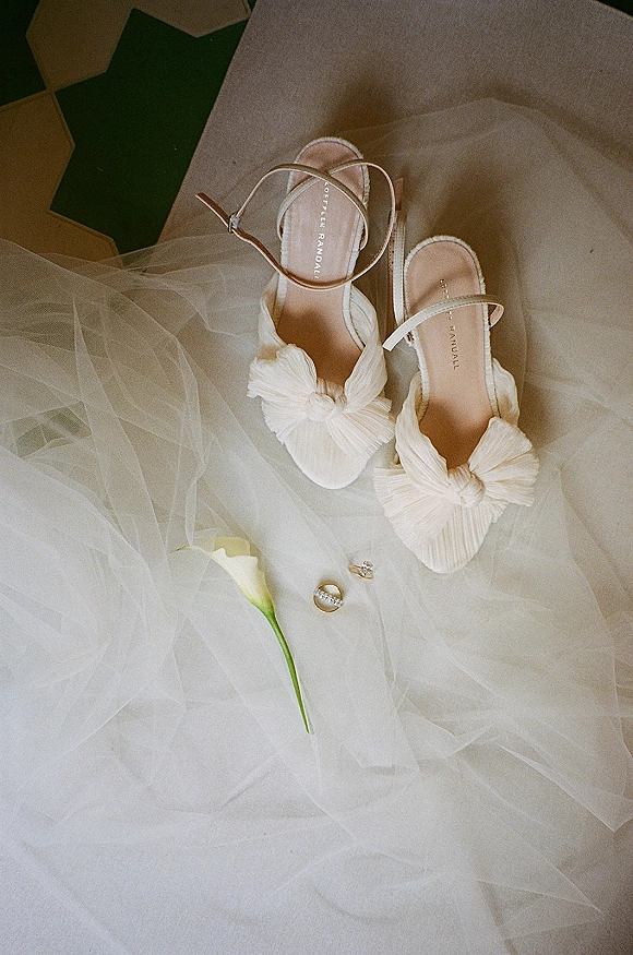 Bridal shoes in an ivory wedding shoe flatlay with bow heels and ankle straps beside wedding rings, tulle fabric, and a calla lily on tile floor