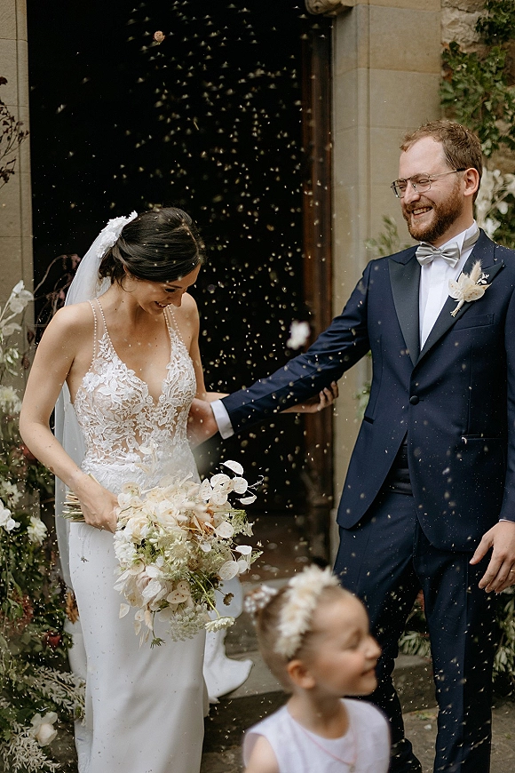 Wedding recessional as bride and groom laugh walking through a confetti toss, veil and bouquet in hand, exiting a stone doorway with florals