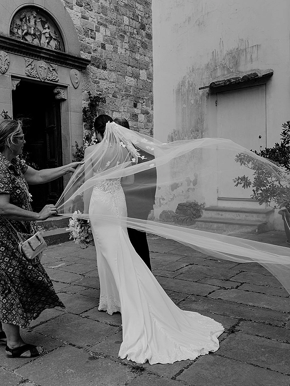 Bride and groom in a long veil wedding photo, walking by an embellished church doorway as her lace dress train flows behind them