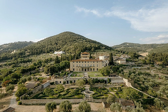 Wedding venue aerial view of a terracotta-roof villa with manicured hedges and a fountain, set among rolling countryside hills under blue sky