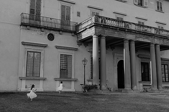 Flower girls running in tulle dresses across a lawn, candidly laughing in front of a historic columned building facade with balcony