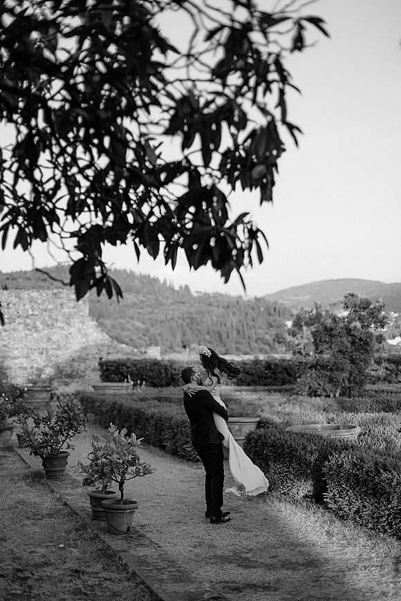 Wedding kiss portrait of groom lifting the bride as they kiss on a garden path, her wedding dress train flowing beside potted plants
