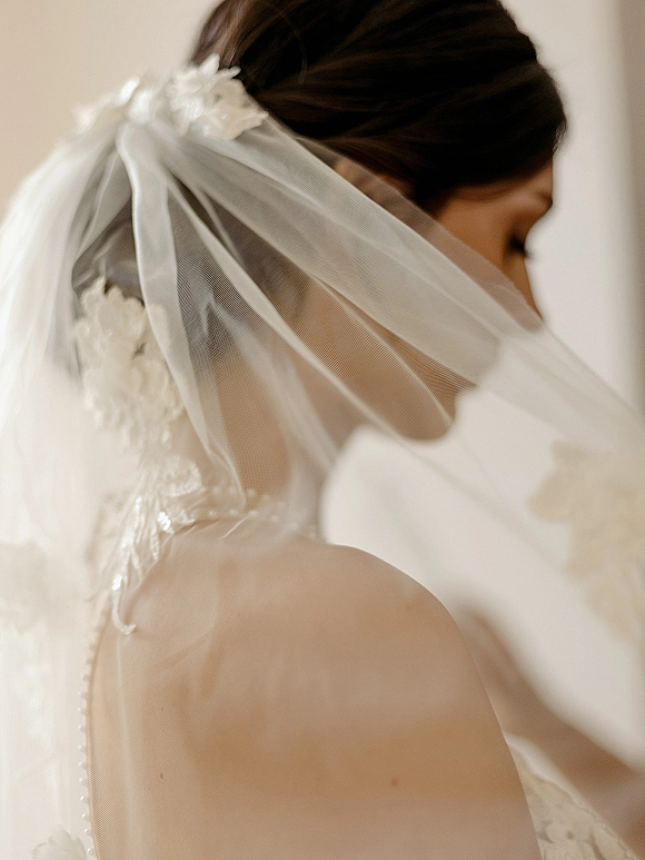 Bridal veil draped over a bride’s updo, showing a floral hairpiece and button-back lace wedding dress against a neutral indoor wall