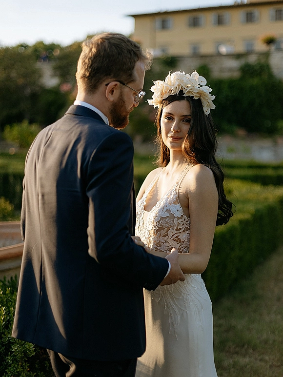 Couple portrait of bride and groom holding hands, her floral crown and lace dress as he looks at her in a villa garden setting
