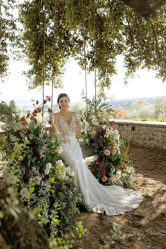 Bridal portrait of a bride on swing in a lace wedding dress with a long train, framed by roses and greenery under a tree canopy outdoors