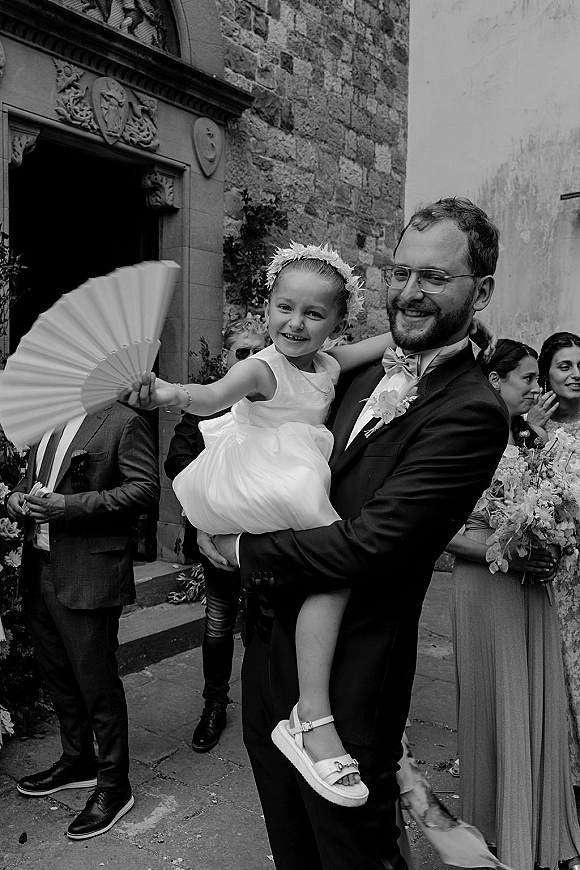 Wedding guest portrait of groom holding a flower girl holding fan, in front of a stone church arched doorway with guests behind