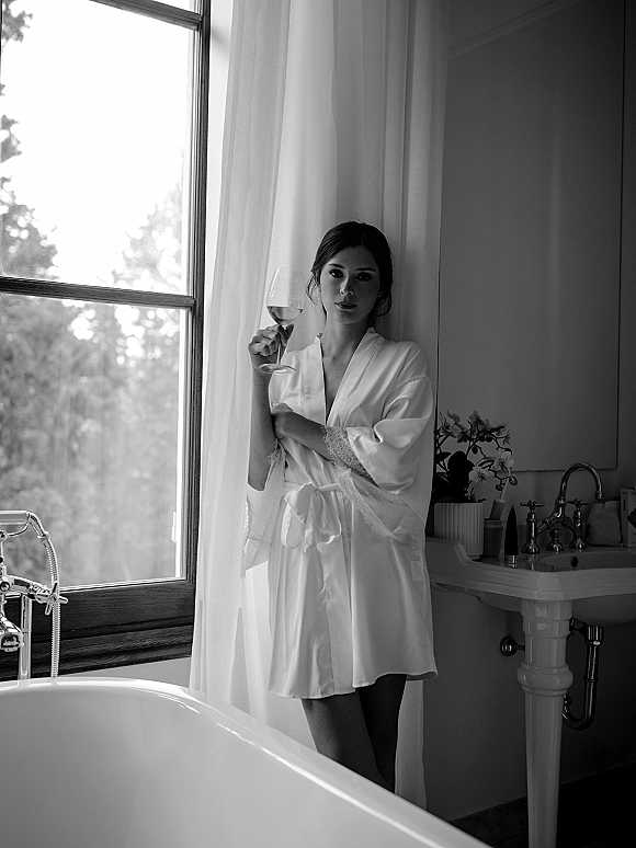 Bridal getting ready as a bride in robe stands by a large window, lace-trimmed satin sash tied, holding a wine glass in a vintage bathroom