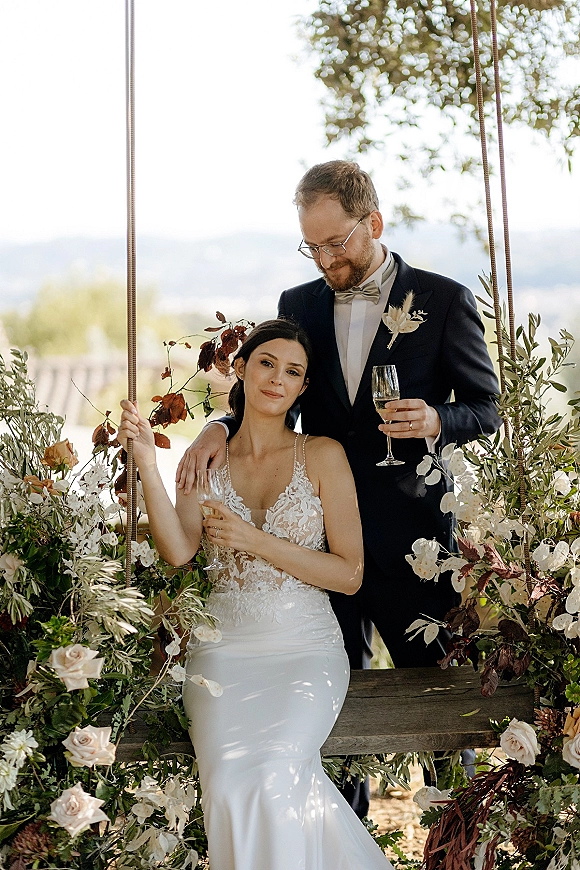 Couple portrait of bride on swing and groom behind her holding champagne flutes, framed by rose greenery with hills and sky beyond
