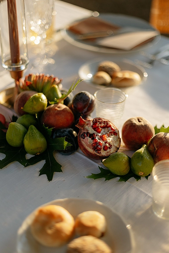Reception tablescape with wedding fruit centerpiece of pomegranates, figs and grapes, taper candles and gold flatware on a sunlit white tablecloth