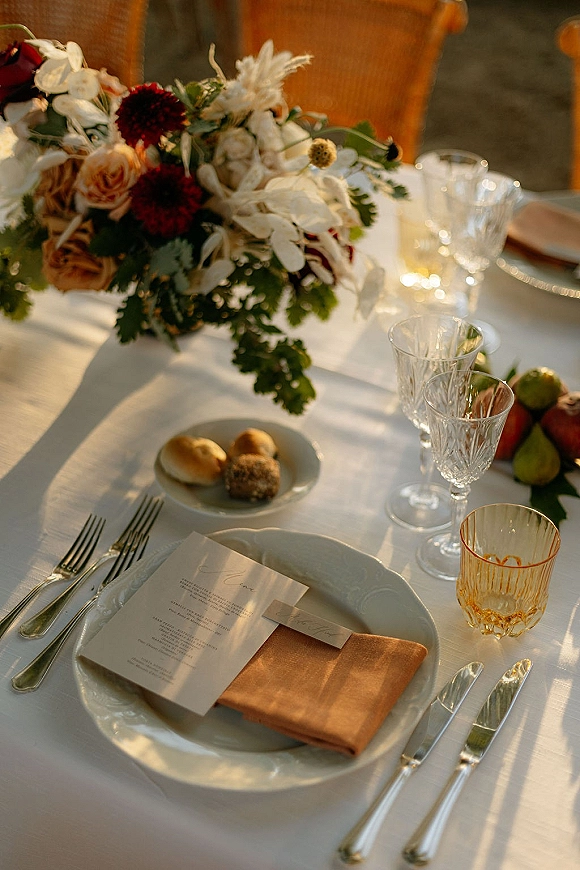 Reception tablescape with wedding table setting, roses and greenery centerpiece, menu and place cards, crystal glasses and amber goblet on sunlit patio