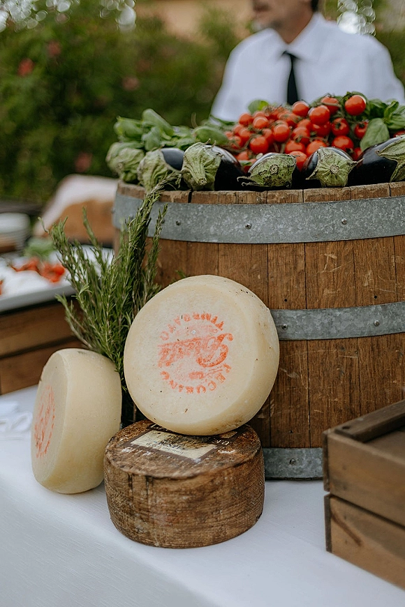 Wedding catering display with rustic wedding food display of cheese wheels, cherry tomatoes, eggplant, and basil on wood crates by greenery outdoors