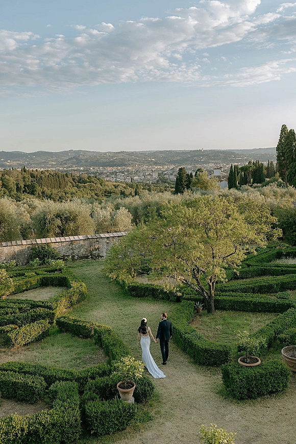 Couple portrait of bride and groom holding hands, walking away in a formal garden maze with a lace wedding dress train and cloudy skyline view