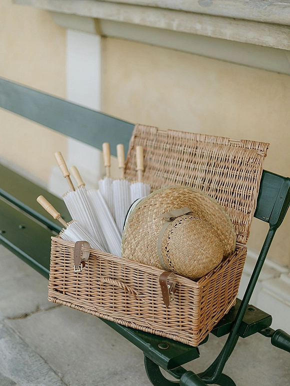 Wedding welcome basket with paper parasols and a straw hat in a wicker basket, set on a green bench by stone steps outdoors