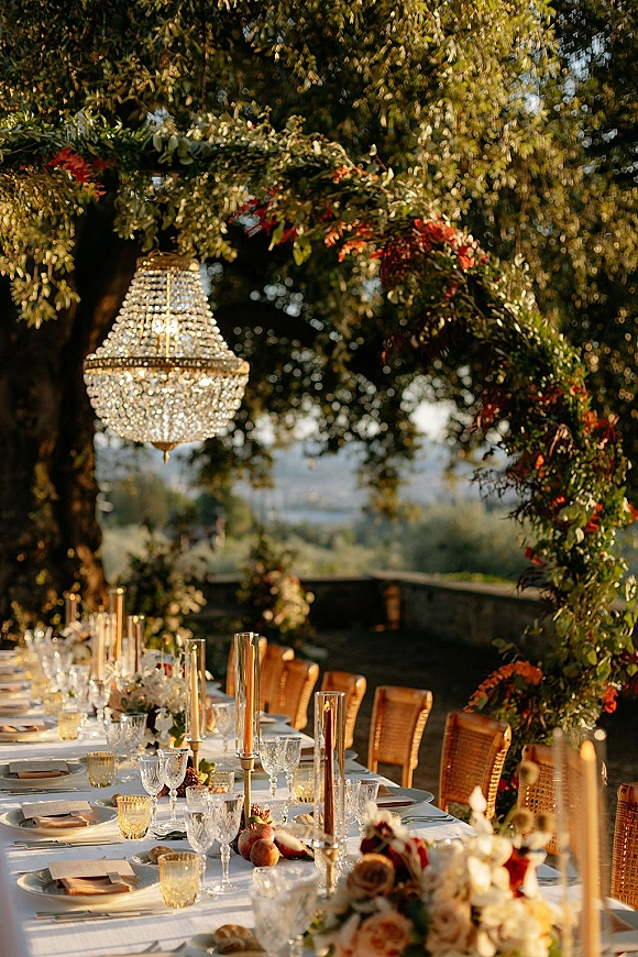 Reception tablescape with a long banquet table wedding setup, taper candles and floral garland under a chandelier in a garden setting